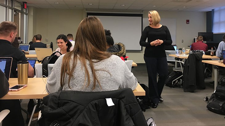 Business professor talks and walks through a classroom while students listen