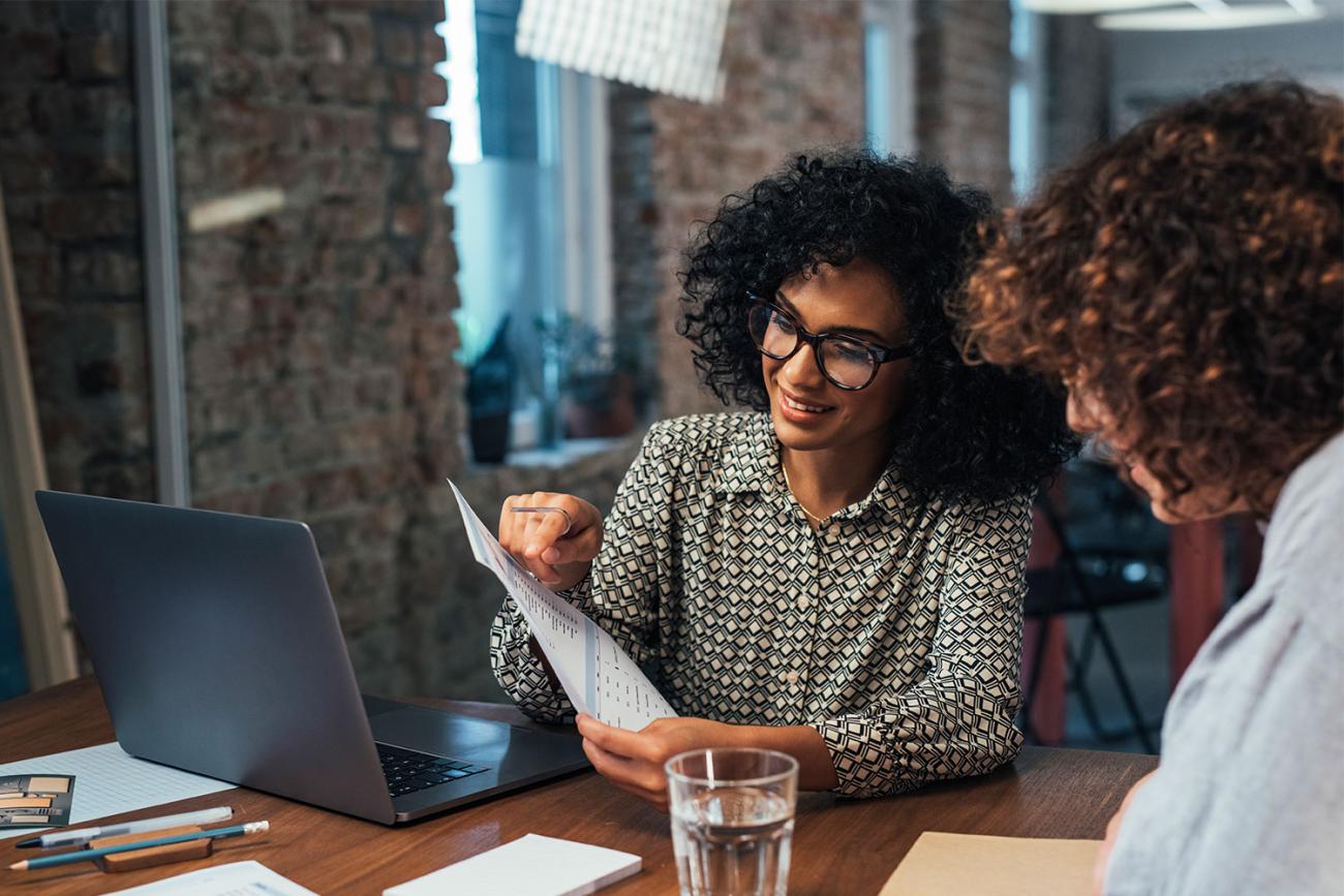 Two women with curly hair sitting at table with laptop looking at a piece of paper one is holding