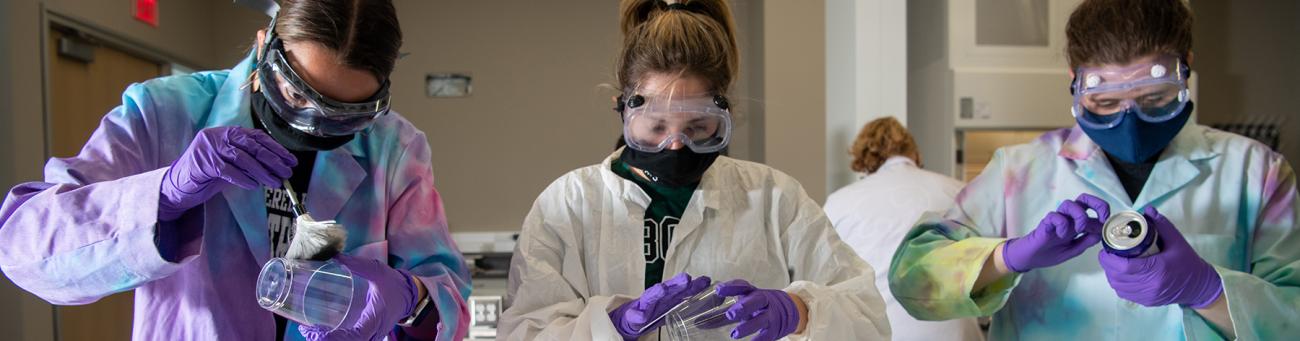 Three chemistry students in a lab with goggles and lab coats on