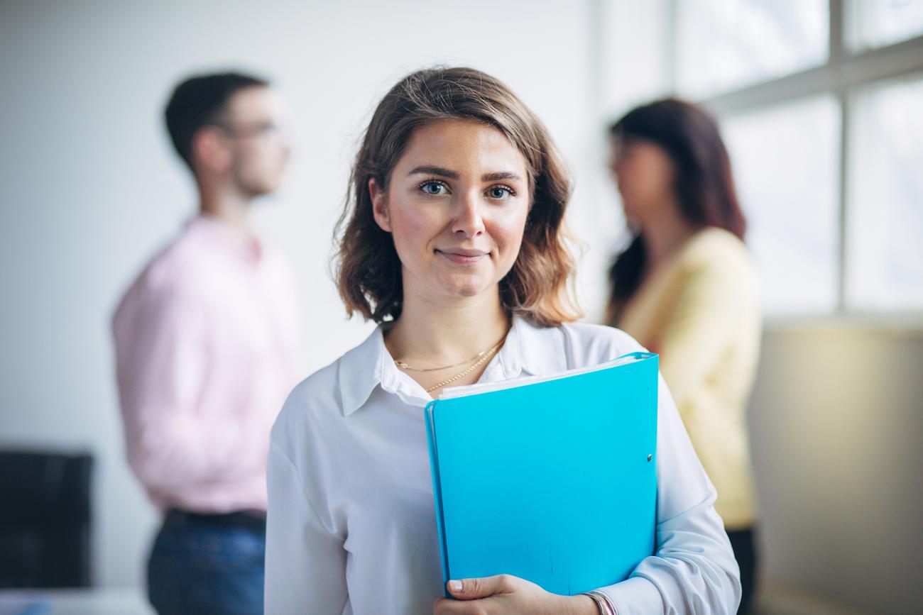 Student looking at the camera holding a book with two people standing and talking behind her