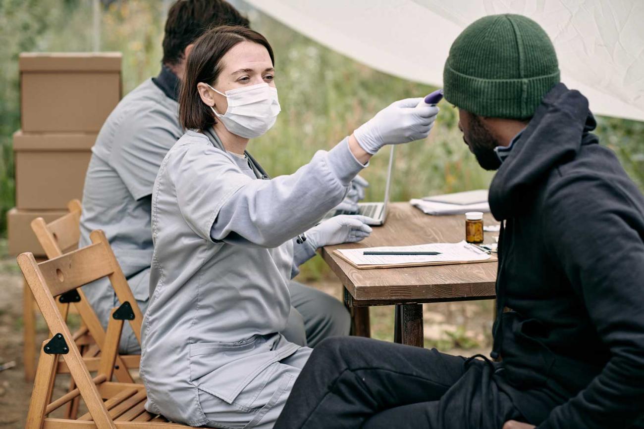 Woman in scrubs sitting at a table outside holding a thermometer to a mans head in front of her