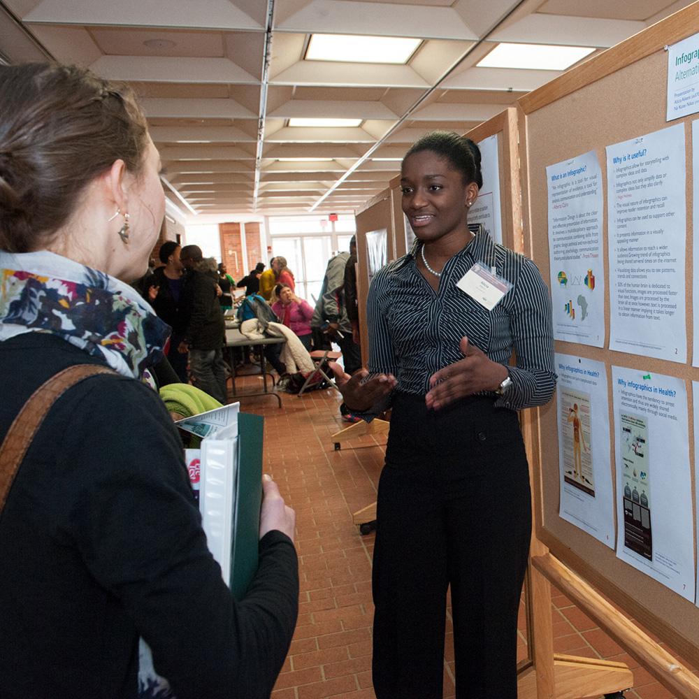 A student shares their work, which is displayed on a posterboard, with another student.