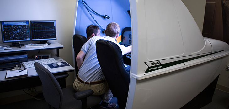 Two people seated in the cockpit of a simulated airplane with opaque front windows and one side of the fuselage cut away