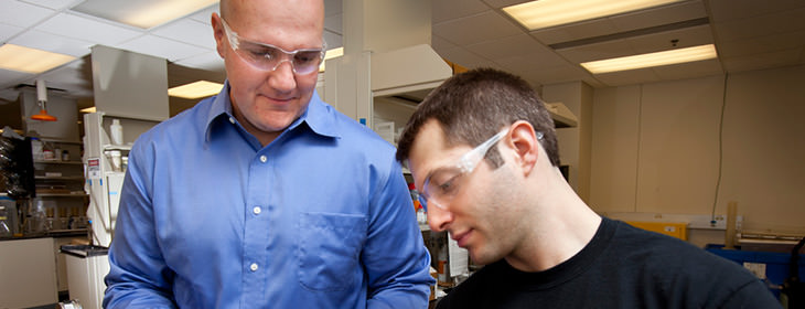 Professor and student wearing safety goggles looking at something on a desk