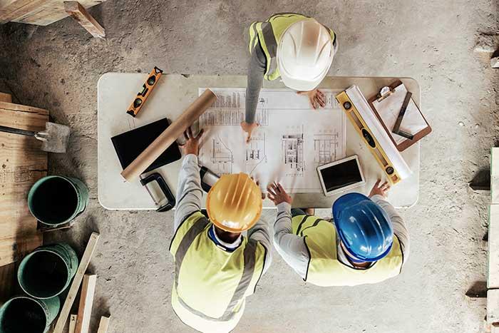 Three construction engineers in hardhats standing over a work table with blueprints