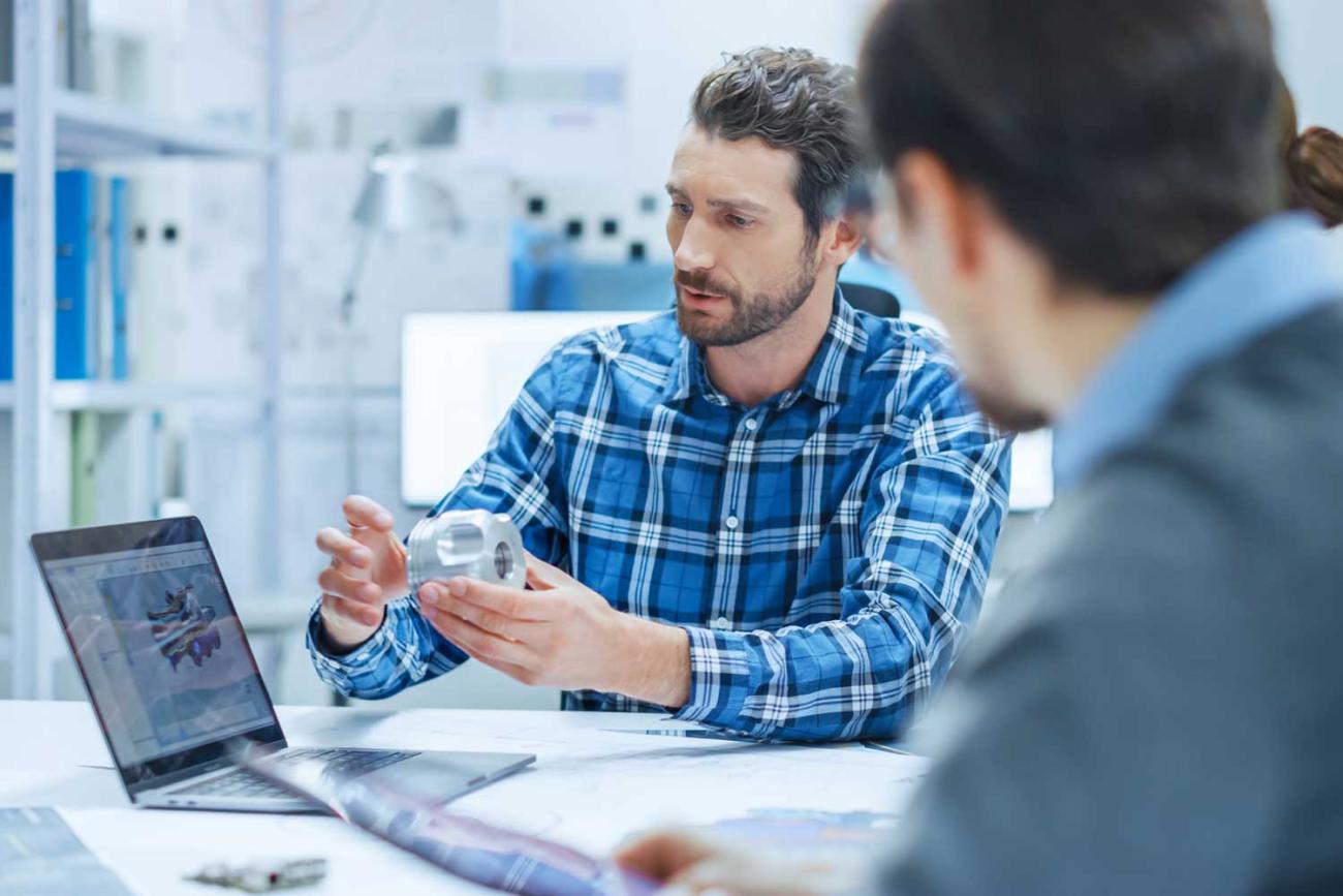 Man in blue shirt holding metal part above table teaching student about engineering analytics