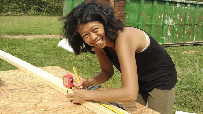 A women smiling and measuring out a piece of building wood with a tape measure. 