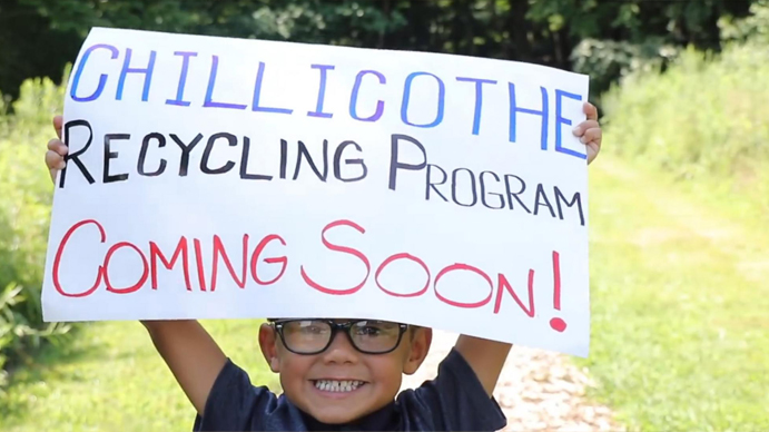 A smiling boy holding a hand written sign saying "Chillicothe Recycling Program Coming Soon"