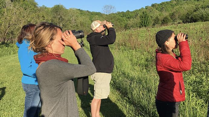 Four people, one man and 3 women standing outside in a green, wooded area looking through binoculars at something in the distance.  