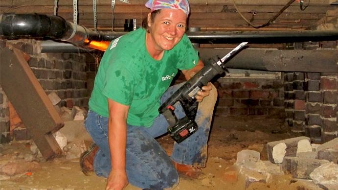A woman holding a power tool is working underneath a house.