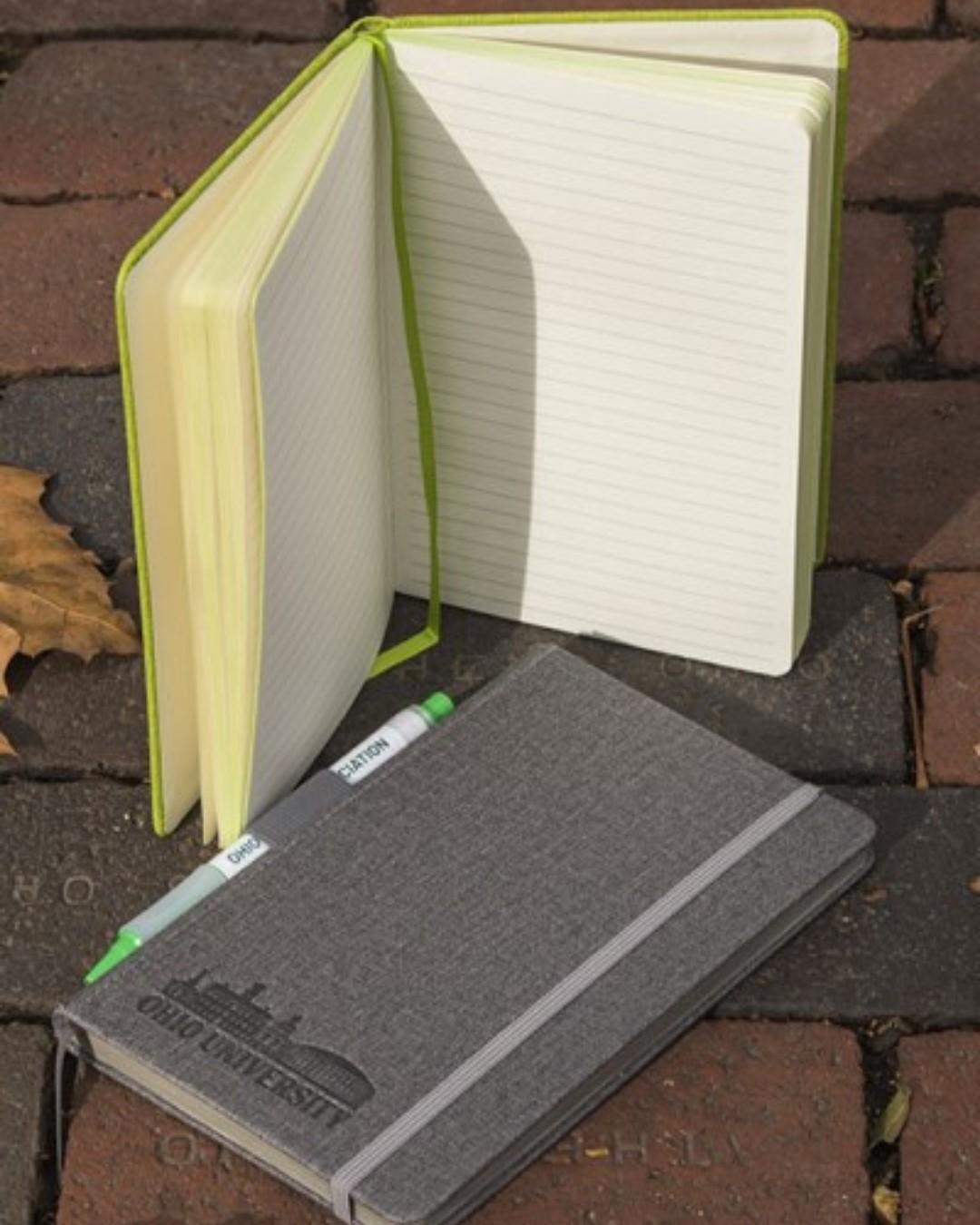 An open green journal and gray journal laying on bricks with iconic Ohio University buildings design.