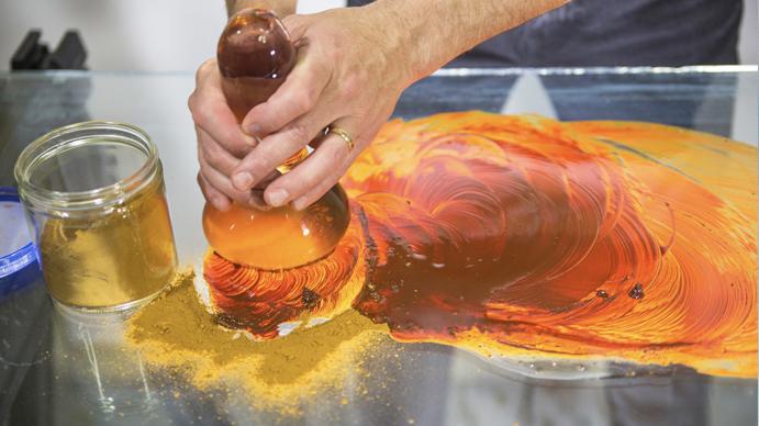 Orange and red powder and paint on metal table with hands using tool to mix.