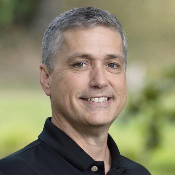 Richard Carr. A man smiles at the camera, he has short gray hair and is wearing a black collared shirt. The background is outdoors with unfocused trees and landscaping.