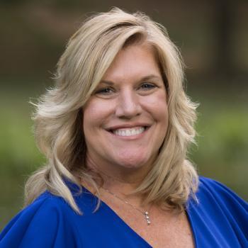 Headshot of Robin Ambrozy. A woman smiles at the camera, she has shoulder length blonde hair, and is wearing a royal blue blouse. The background of the image is unfocused landscape.